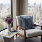 Load image into Gallery viewer, A modern upholstered armchair with a textured light beige seat and back, a dark wood frame, and a navy blue patterned throw pillow, positioned in a bright room with a city view from a large window. A small marble side table next to the chair holds a vase of purple flowers and a framed picture.
