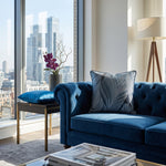 Load image into Gallery viewer, A luxury blue velvet tufted sofa in a modern loft apartment with a city skyline view, styled with a blue accent pillow and a gray patterned throw pillow.

