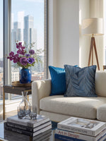 Load image into Gallery viewer, A modern living room corner with a cream textured sofa, two blue throw pillows, a side table with a blue vase of purple flowers, and a large window showing a city skyline. Books and glassware sit on a glass table in the foreground.
