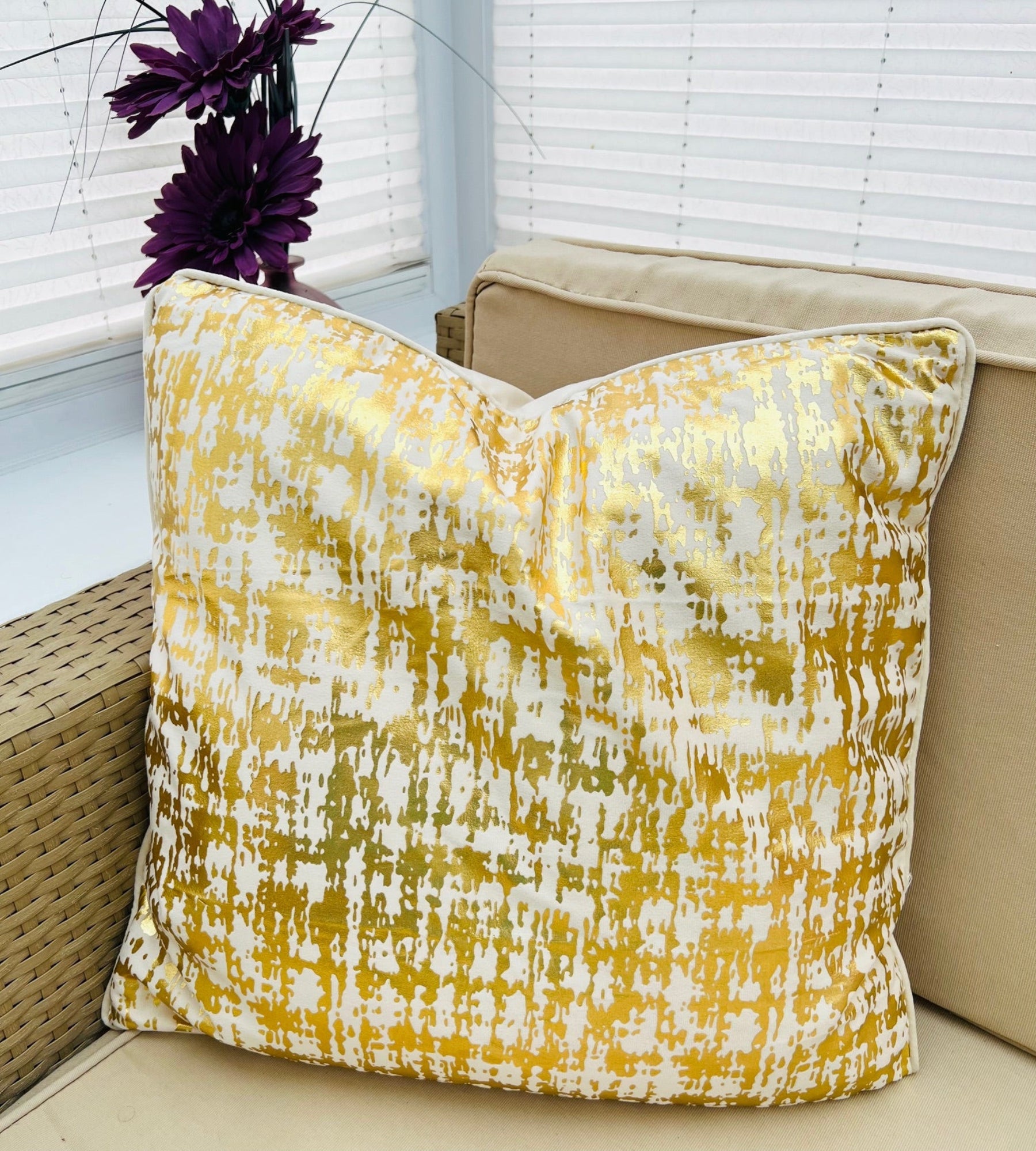 Gold and beige patterned pillow on a beige couch with purple flowers in the background.