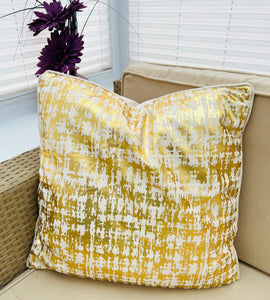 Gold and beige patterned pillow on a beige couch with purple flowers in the background.