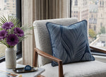 Load image into Gallery viewer, A textured light gray armchair in a bright room with a window, featuring a navy blue throw pillow with a white leaf pattern. A side table with purple flowers and a small blue object sits beside the chair.
